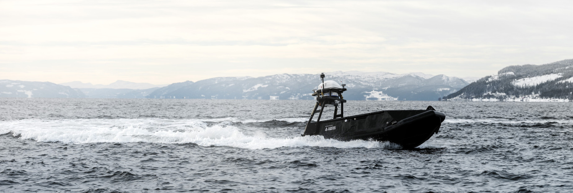 Side profile of the Mariner USV navigating nearshore waters in a fjord with snowy mountain backdrop.